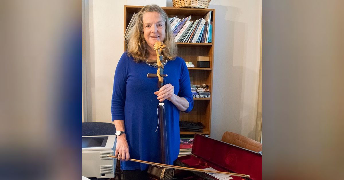 Penny Anderson Brill wearing a blue shirt stands with a cello