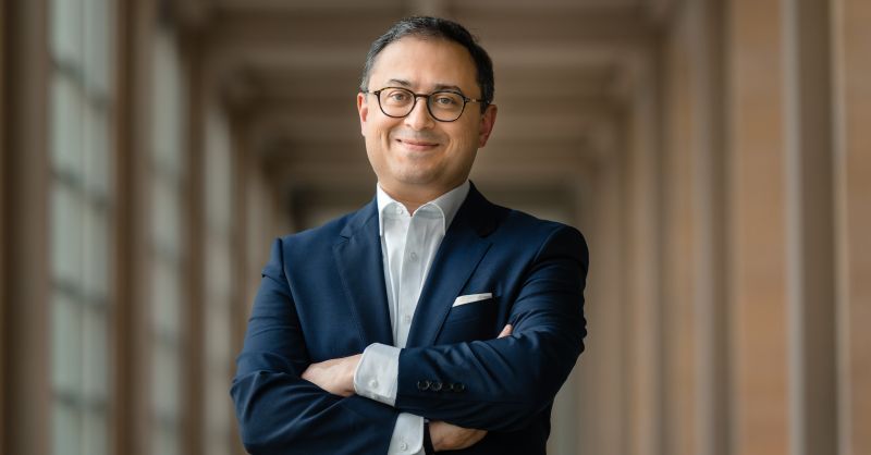 John Turman in a suit and glasses stands confidently in a well-lit hallway