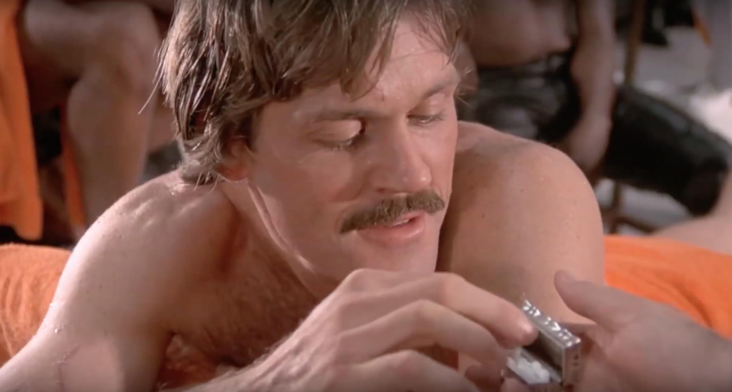 John R. Beck with a mustache and a visible tattoo lies on a bed